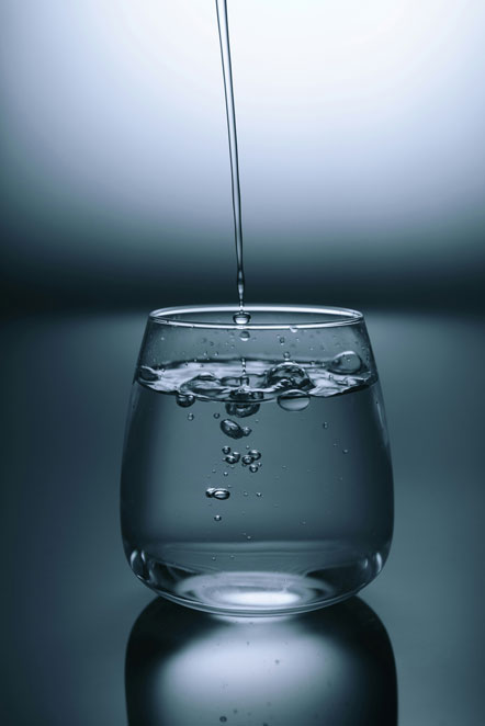 Clear drinking glass with water spilling in on a gray surface